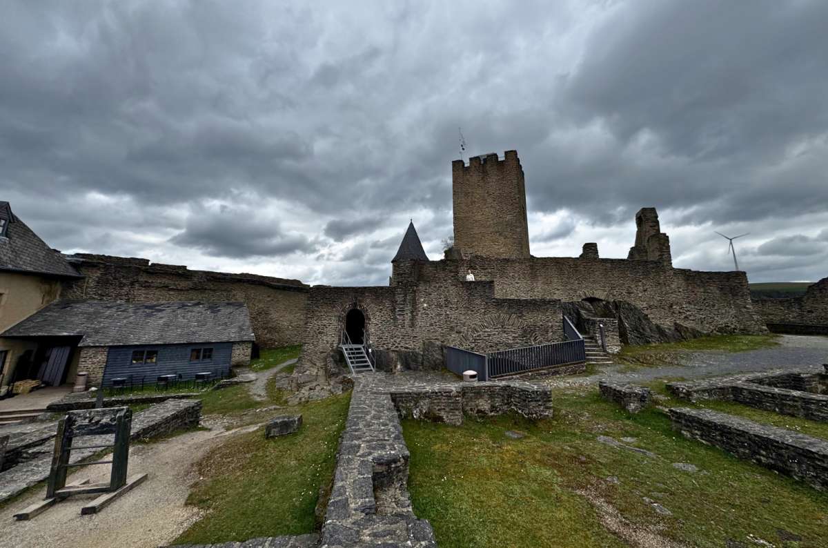 a photo of the courtyard of Bourscheid Castle ruins, showing the keep, arched gateway, and surrounding stone walls under a cloudy sky, Luxembourg, photo by Next Level of Travel