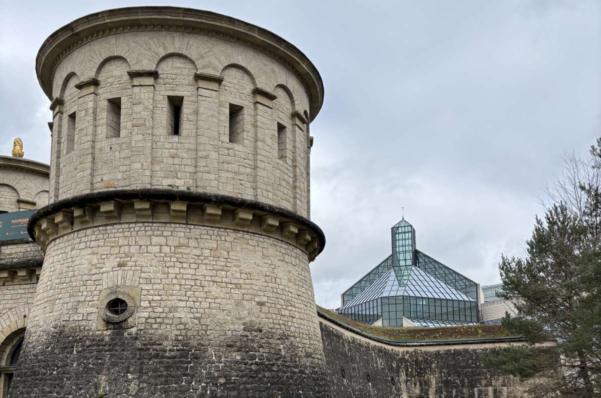 a photo of the cylindrical stone tower of Musée Dräi Eechelen with the glass pyramid of MUDAM visible behind it, Kirchberg, Luxembourg City, photo by Next Level of Travel