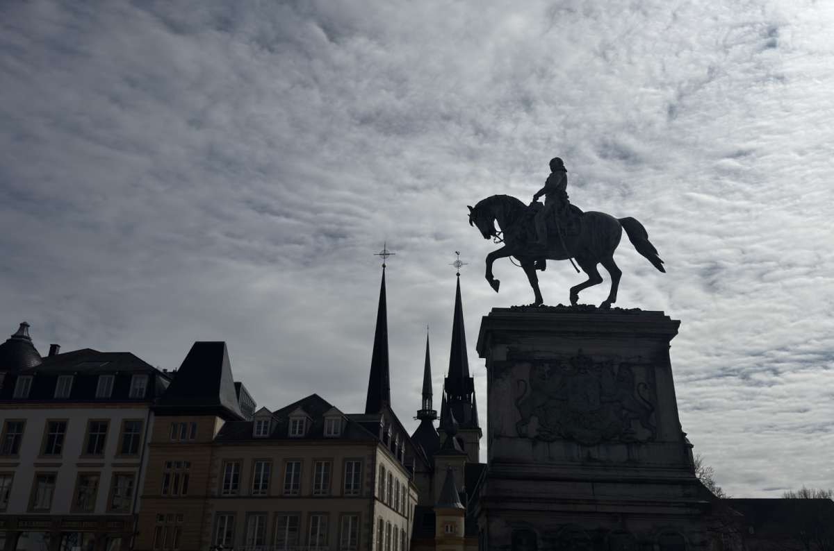 a photo of the equestrian statue at Place Guillaume II, with Notre-Dame Cathedral spires visible in the background, Luxembourg, photo by Next Level of Travel