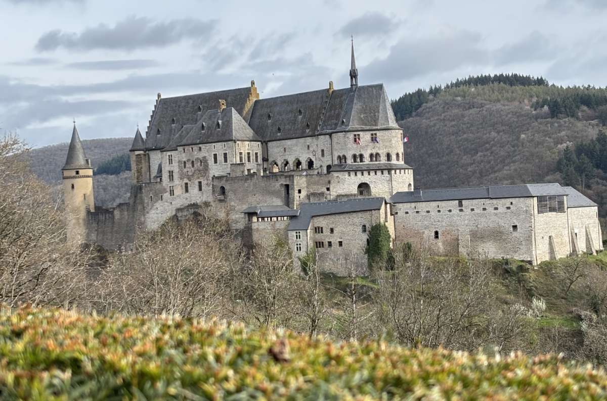 a photo of Vianden Castle with its towers, slate rooflines, and layered stone walls rising above bare spring trees and hedgerows, Luxembourg, photo by Next Level of Travel
