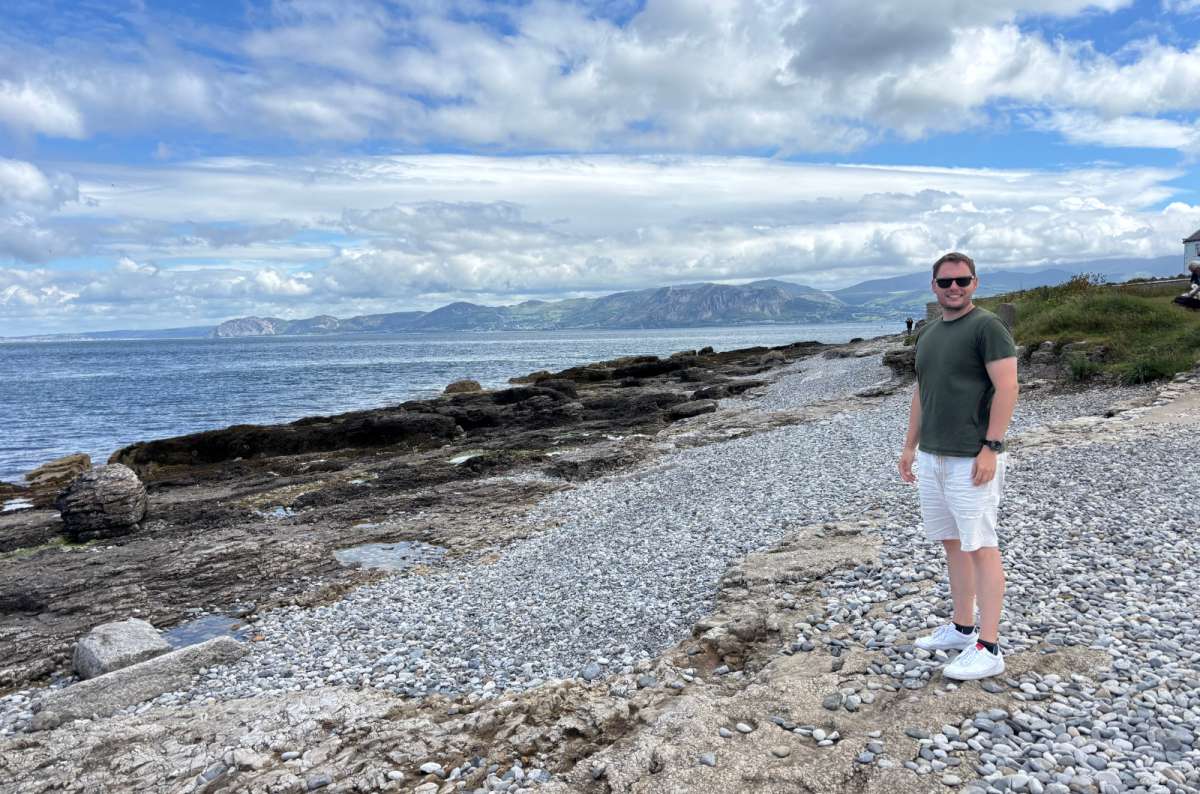 a photo of a tourist on a pebble beach on Anglesey with the mainland headlands across the water, Anglesey, Wales, photo by Next Level of Travel
