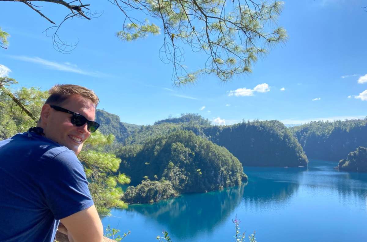 a photo of a smiling tourist looking back over his shoulder above a blue lake surrounded by forested hills, Lagunas de Montebello, Mexico, photo by Next Level of Travel
