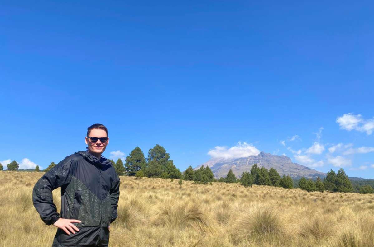 a photo of a tourist in a black windbreaker standing in a golden grass meadow with Iztaccíhuatl in the background, Paso de Cortés, Izta Popo National Park, Mexico, photo by Next Level of Travel