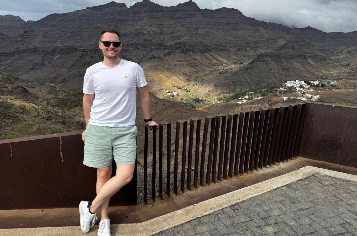 a photo of a tourist posing in front of a railing at a hike, Mirador Veneguera visible in the background, Canary Islands, photo by Next Level of Travel
