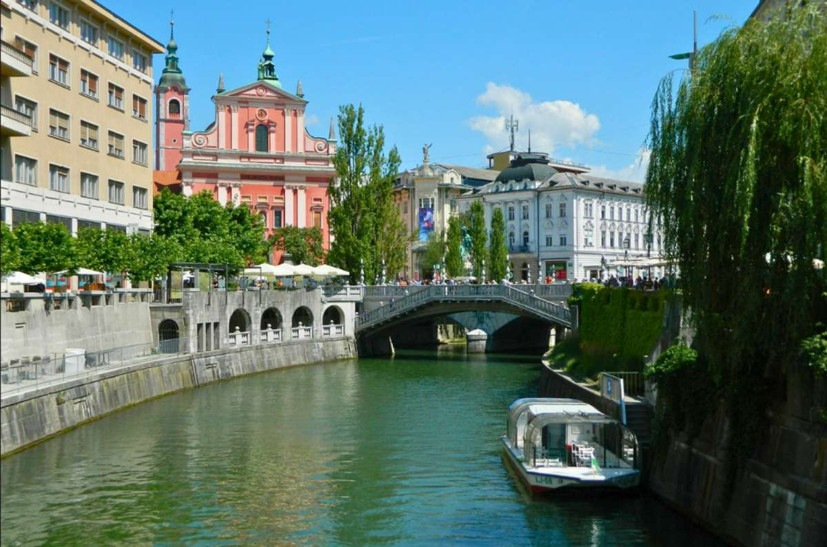 a photo of a tourist boat on the Ljubljanica River with the Triple Bridge and the Franciscan Church in the background, Ljubljana, Slovenia