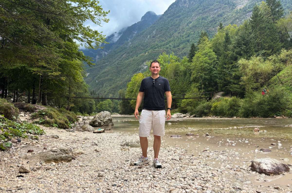 a photo of a tourist standing on a pebble riverbank of the Soča River with a hanging footbridge and forested mountains behind, near the Great Soča Gorge, Slovenia, photo by Next Level of Travel