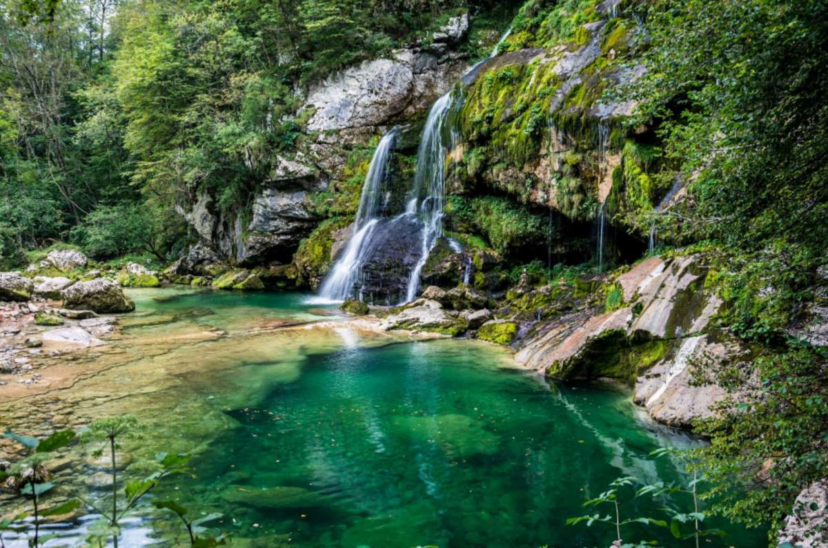 a photo of Virje Waterfall falling into a green pool surrounded by mossy rocks, near Bovec, Slovenia