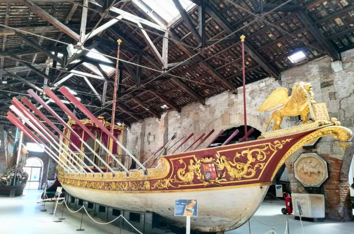 a photo of a gilded ceremonial rowing barge with a golden winged lion of St. Mark on its bow in the Ships Pavilion of the Naval History Museum, Venice, Italy, photo by Next Level of Travel