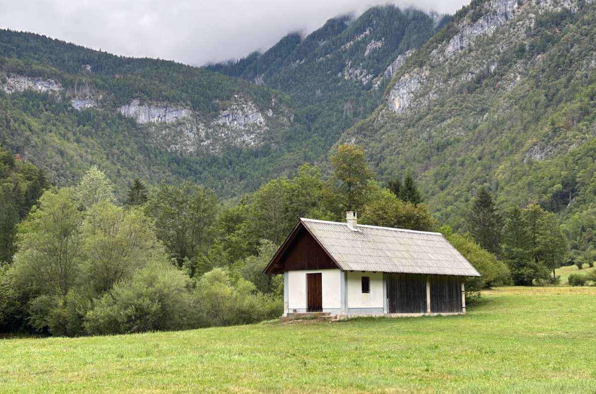 a photo of a white herdsman's hut in the green meadows of the Voje Valley below forested mountains, Slovenia, photo by Next Level of Travel