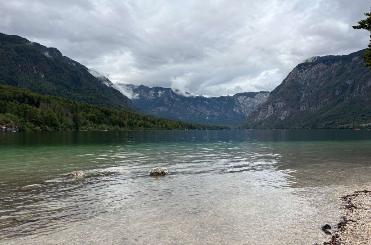 a photo of calm crystal-clear water by a pebble shore with Lake Bohinj and the mountains behind, Slovenia, photo by Next Level of Travel