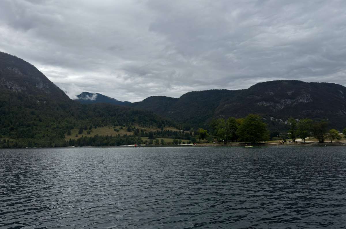 a photo of dark water and the forested northern shore of Lake Bohinj under heavy grey clouds, Slovenia, photo by Next Level of Travel