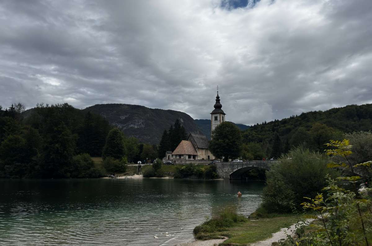 a photo of the Church of St. John the Baptist and the stone bridge at the eastern end of Lake Bohinj under dark clouds, Ribčev Laz, Slovenia, photo by Next Level of Travel