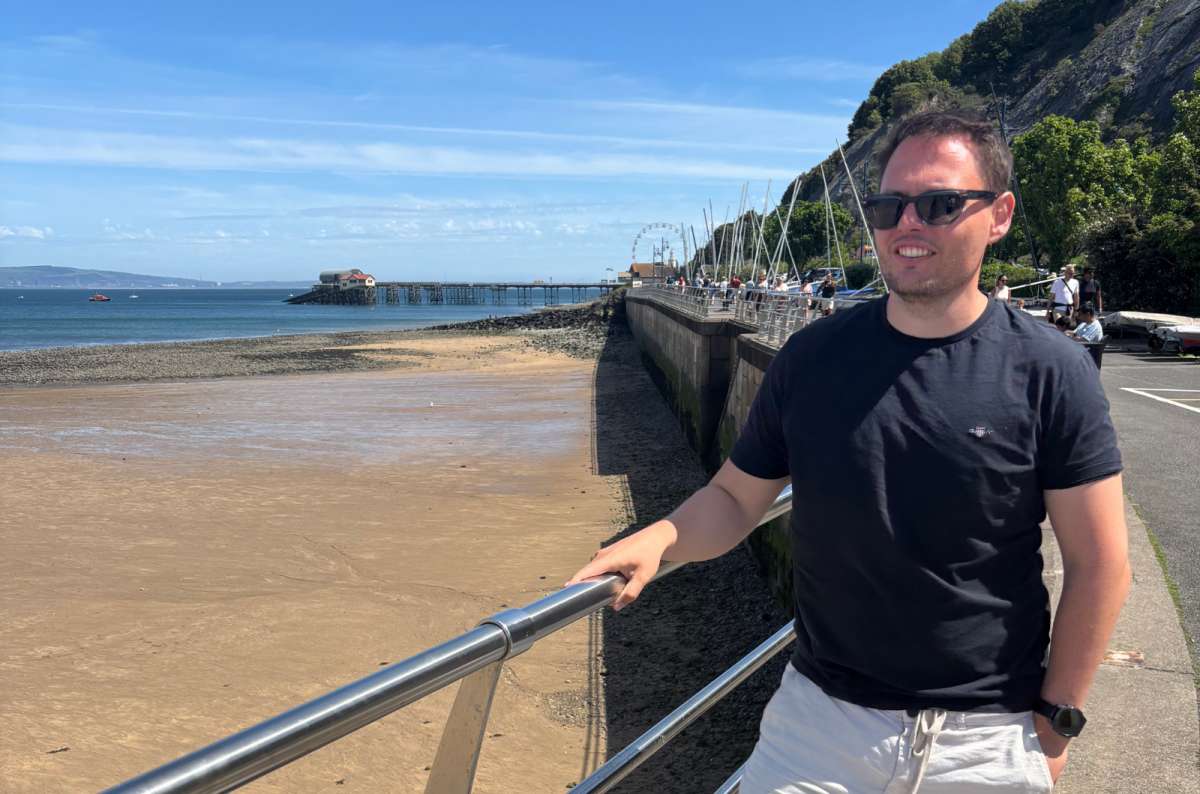 a photo of a tourist leaning on the promenade railing above the sands of Swansea Bay with Mumbles Pier in the distance, Wales, photo by Next Level of Travel