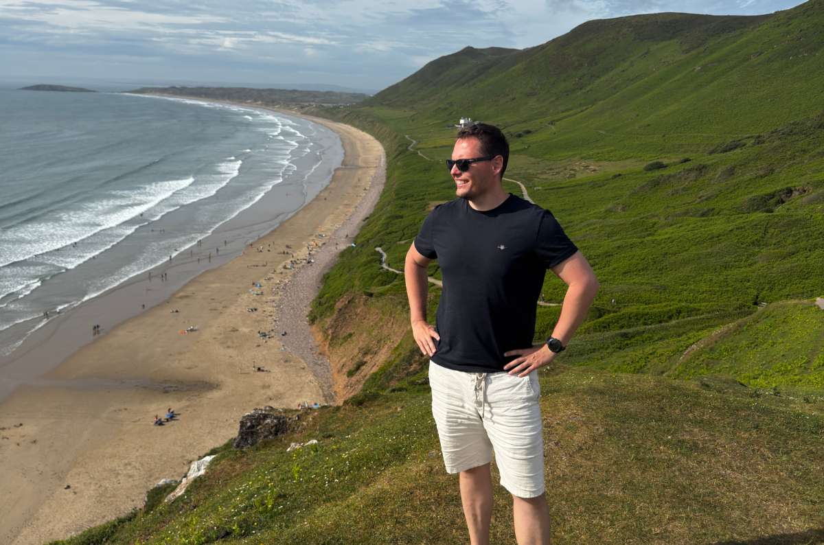 a photo of a tourist on the clifftop above Rhossili Bay with the beach curving away below, Wales, photo by Next Level of Travel