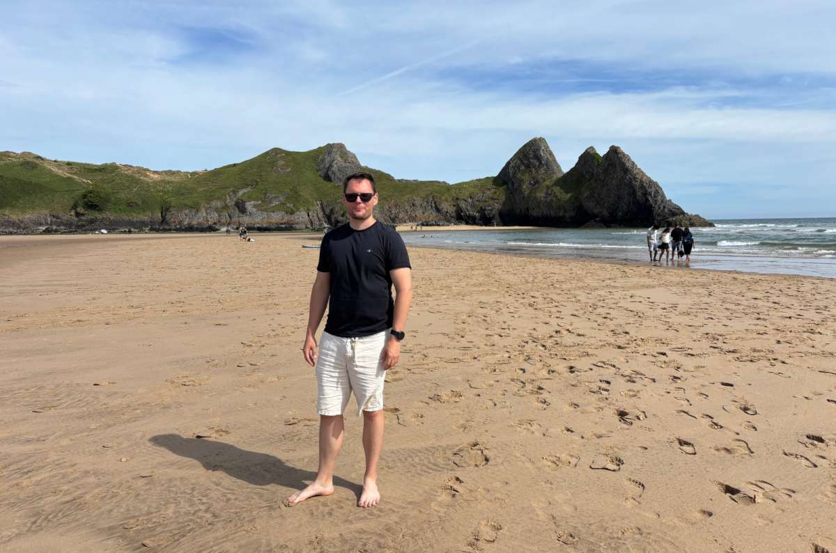 a photo of a tourist standing barefoot on the golden sand of Three Cliffs Bay with the three limestone cliffs behind, Wales, photo by Next Level of Travel