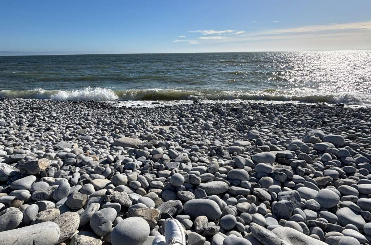 a photo of a tourist's feet resting on the large grey pebbles of Dunraven Bay beside the sea, Wales, photo by Next Level of Travel