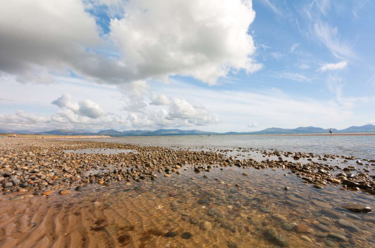 a photo of pebbles and shallow water at Llanddwyn Beach with the mountains of Eryri on the horizon, Anglesey, Wales