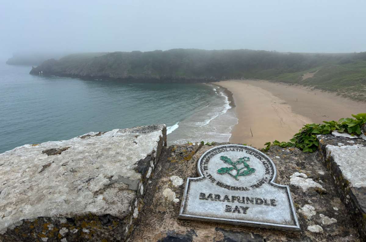a photo of the Barafundle Bay sign on the stone wall above the sandy beach and green cliffs, Wales, photo by Next Level of Travel