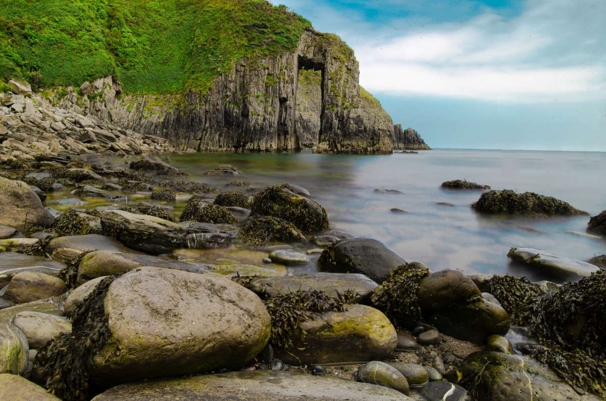 a photo of the natural rock arch of Church Door Cove above the boulders at low tide, Pembrokeshire, Wales