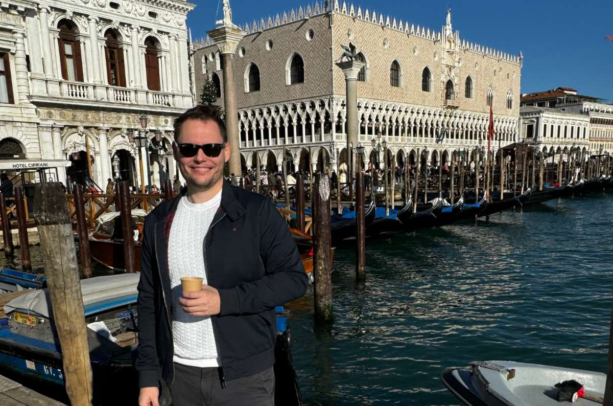 a photo of a tourist standing on a wooden pier by the Venetian lagoon with gondolas and the Doge's Palace behind, Venice, Italy, photo by Next Level of Travel