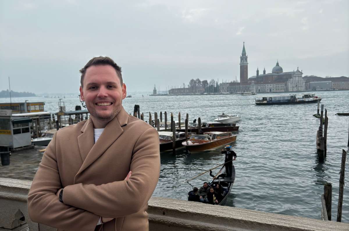 a photo of a tourist on the San Marco waterfront with a gondola, water taxis and San Giorgio Maggiore across the lagoon, Venice, Italy, photo by Next Level of Travel
