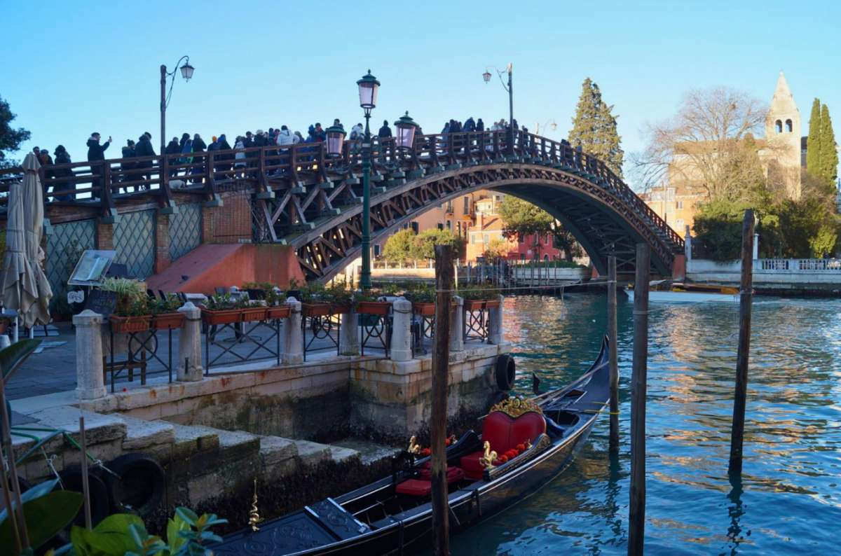 a photo of the wooden Ponte dell’Accademia crowded with tourists over the Grand Canal with a gondola moored below, Venice, Italy