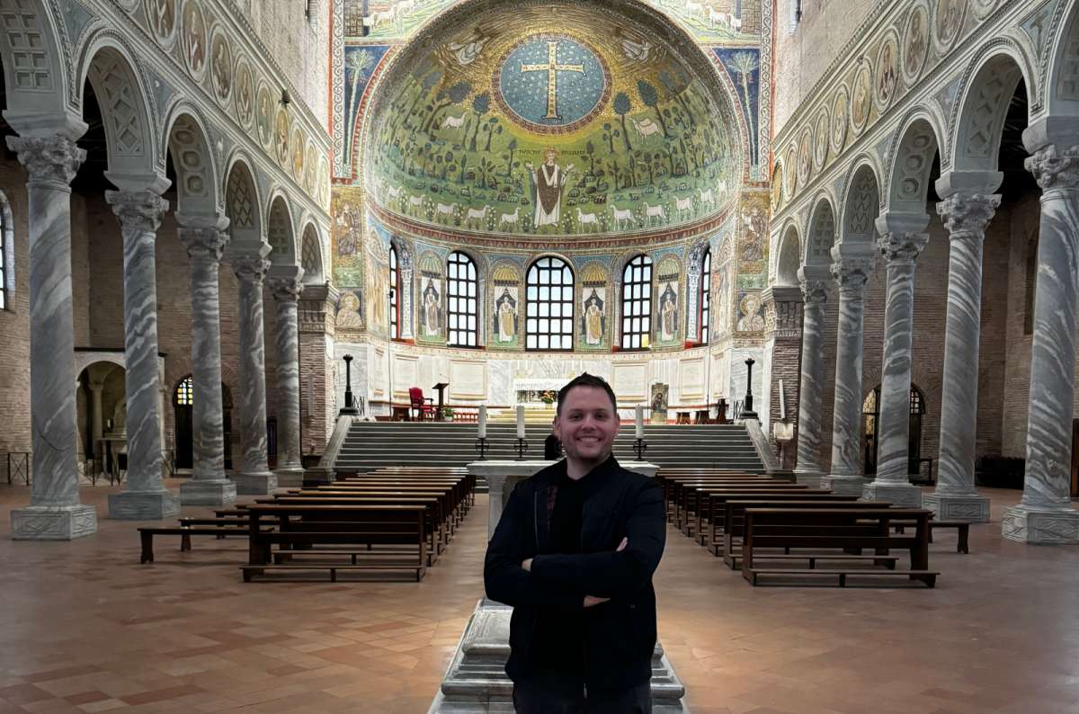a photo of a tourist standing in the nave of the Basilica of Sant'Apollinare in Classe with the green and gold apse mosaic behind, Ravenna, Italy, photo by Next Level of Travel