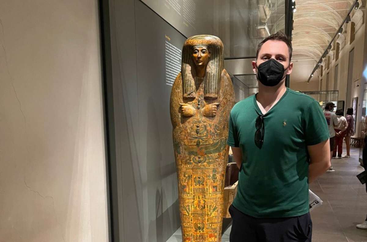 a photo of a tourist standing next to a painted ancient Egyptian coffin in a glass case in the Egyptian Museum, Turin, Italy, photo by Next Level of Travel