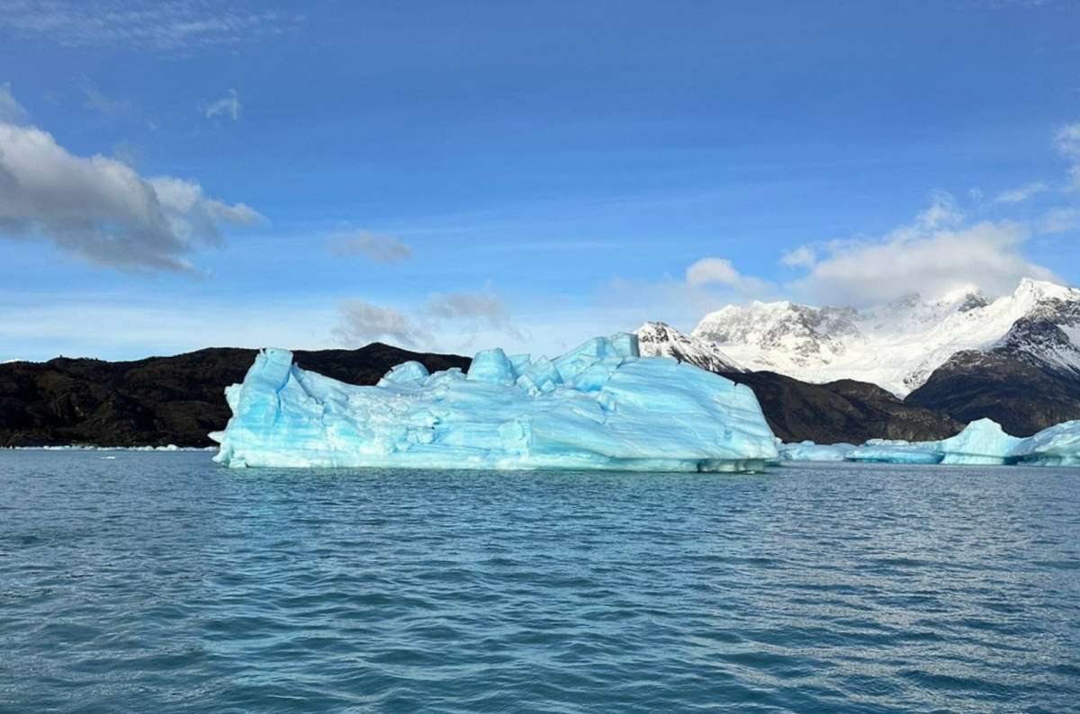 a photo of a large blue iceberg floating on Lago Argentino below snowy mountains, Los Glaciares National Park, Argentina, photo by Next Level of Travel