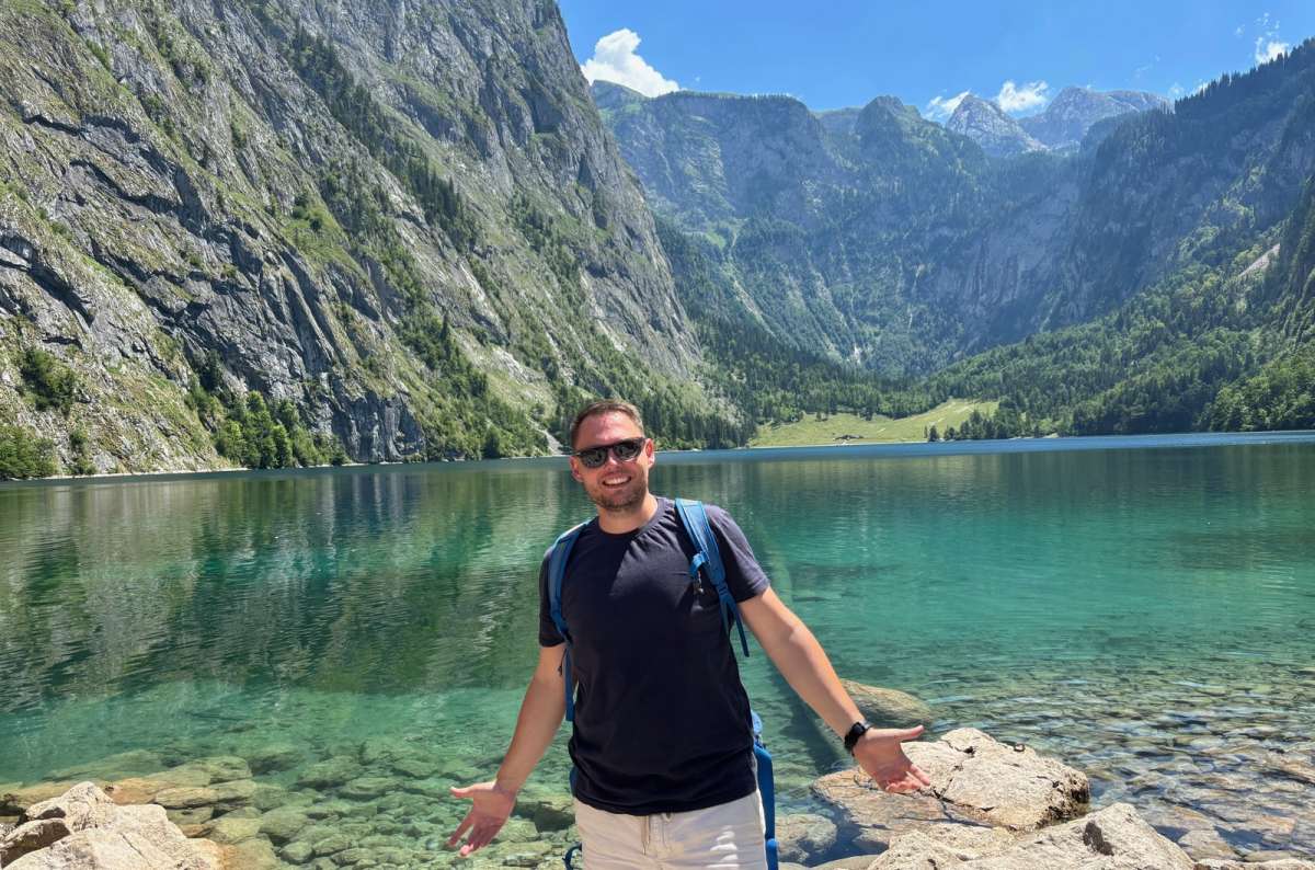a photo of a tourist standing on the rocky shore of Obersee with the Fischunkelalm meadow and mountains behind, Berchtesgaden National Park, Germany, photo by Next Level of Travel
